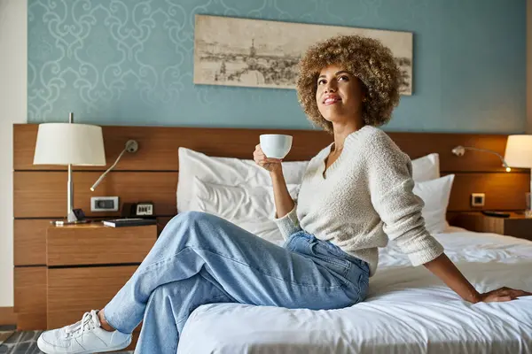 stock image Content and young african american woman enjoying coffee while sitting on hotel bed, weekend getaway