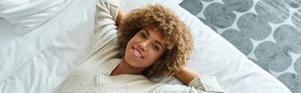 stock image banner of relaxed african american woman smiling and lying on bed in hotel room, solo traveler