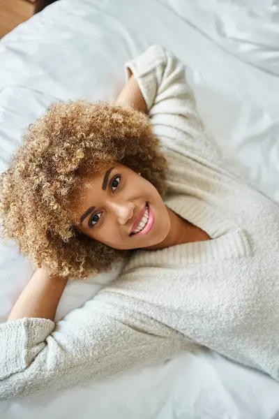 リラックスしたアフリカ系アメリカ人女性のトップビュー笑顔とホテルの部屋のベッドに横たわる — ストック写真