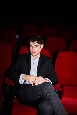 good looking young male model with dapper style posing on red cinema chair and looking away clipart