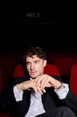 Siyah şık takım elbiseli yakışıklı genç adam sinemada kırmızı sandalyelerde film izliyor, elleri yüzüne yakın.