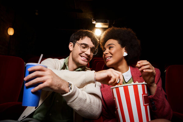 cheerful young multicultural couple in retro clothes enjoying date at cinema and sharing popcorn