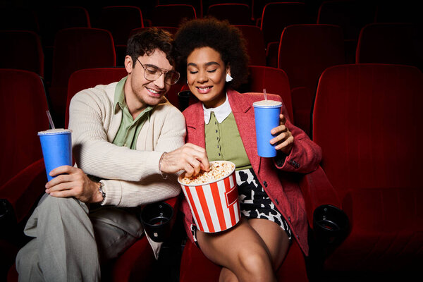 good looking cheerful diverse couple in stylish attires sharing popcorn at cinema, Valentines day
