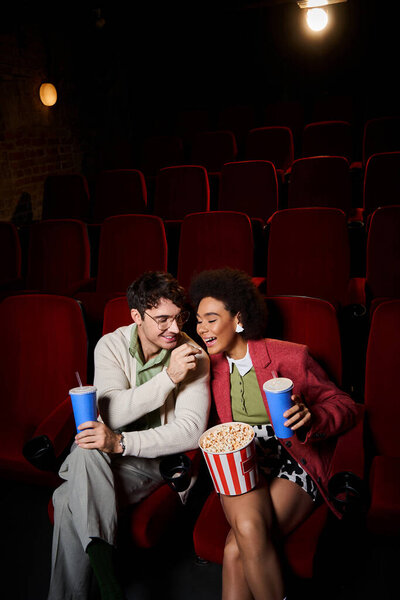 handsome man in glasses sharing popcorn with his african american girlfriend at cinema on date