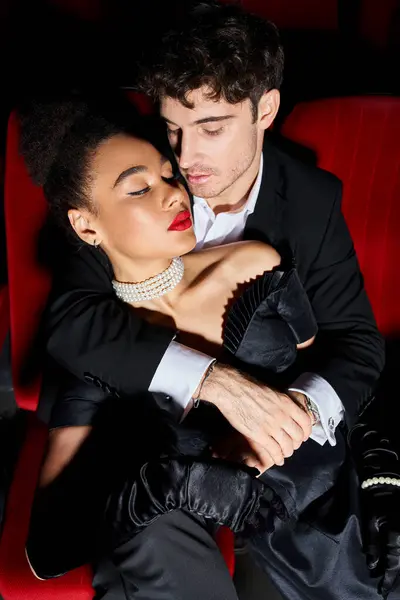 appealing multicultural couple in elegant chic outfits hugging on red cinema chairs while on date
