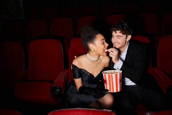 stock image joyful handsome man in black suit feeding popcorn to his african american young girlfriend on date