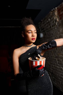 beautiful african american woman in black dress with red lipstick holding popcorn and looking away clipart