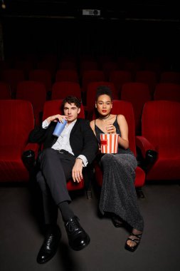 Kırmızı koltuklarda oturmuş gazozlu ve patlamış mısırlı zarif elbiseli çok kültürlü bir çift.