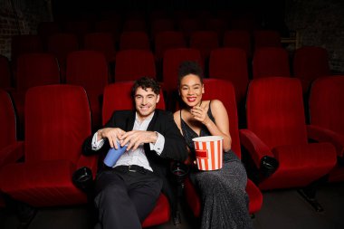 Siyah elbiseli, gazoz ve patlamış mısır taşıyan ve neşeyle gülümseyen neşeli ve çekici bir çift.