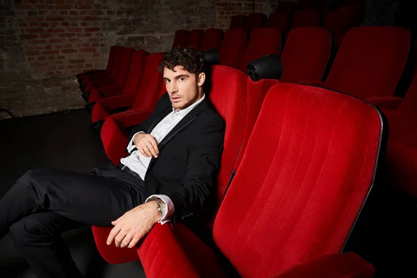 Stock image handsome appealing man in elegant black suit sitting on red chairs at cinema and looking at camera