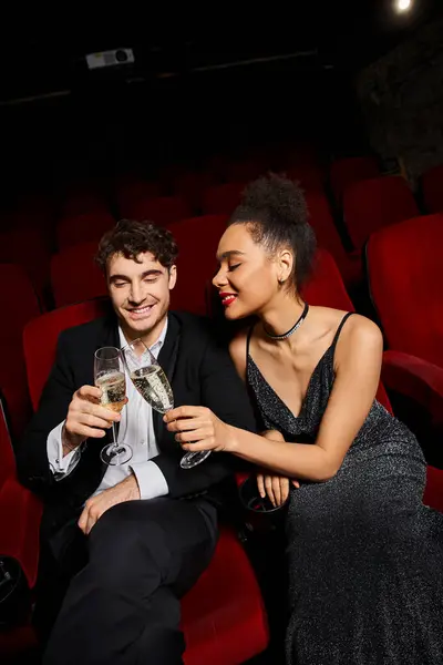cheerful young multicultural couple in elegant outfits clinking glasses while on date at cinema