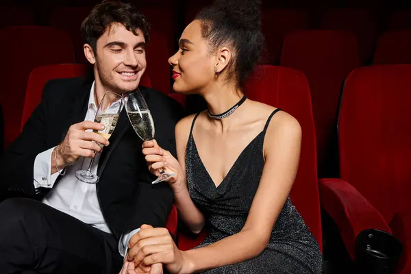 Şık siyah elbiseli çift şampanya bardaklarını tokuşturup birbirlerine bakıyorlar.