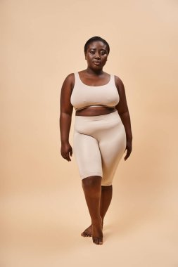 Graceful plus size african american model in beige attire posing against a matching backdrop clipart