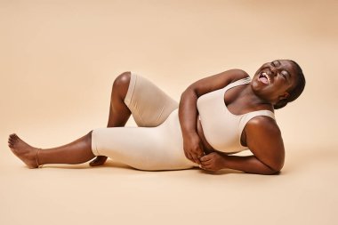 plus size african american woman in beige lingerie laughing and reclining against matching backdrop clipart