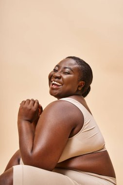 plus size african american model in beige lingerie laughing and reclining against matching backdrop clipart
