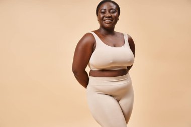 cheerful plus size woman in beige underwear posing in studio, body positive and self esteem clipart