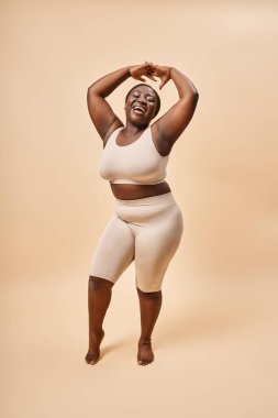 happy plus size woman in beige underwear posing with raised hands, body positive and self esteem clipart