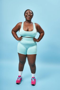 jolly plus size african american woman in blue sportswear with hands on hips on matching backdrop clipart