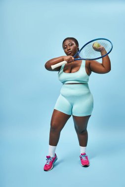 Kendine güvenen Afrikalı Amerikalı bir kadın. Mavi arka planda tenis oynamaya hazır. Vücudu pozitif.