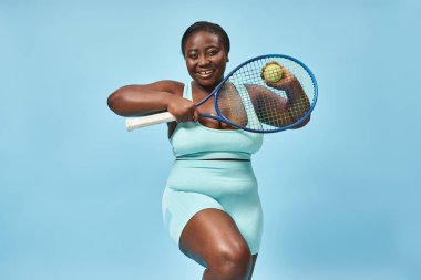 Mutlu artı Afrikalı Amerikalı kadın mavi arka planda tenis oynamaya hazır, vücut pozitifliği.