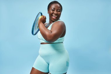 Neşeli ve iri yarı Afrikalı Amerikalı kadın mavi arka planda tenis oynamaya hazır, vücut pozitifliği.