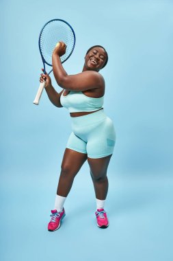 Artı boy Afrikalı Amerikalı kadın mavi arka planda tenis oynamaya hazır, vücut pozitifliği ve spor