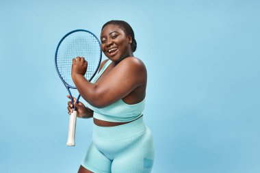 Gülümseme artı boy Afrikalı Amerikalı kadın mavi arka planda tenis oynamaya hazır, vücut pozitifliği.