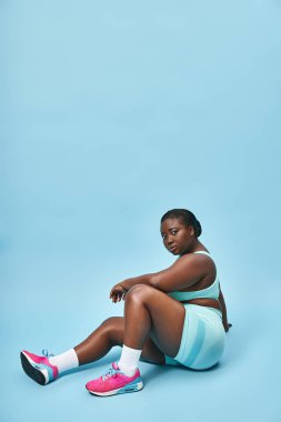 Rahatlamış ve iri yarı Afro-Amerikalı bir kadın mavi spor kıyafetleriyle arka planda oturuyor.