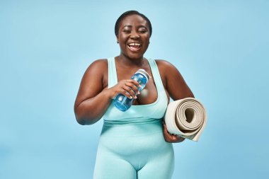 Gülümseyen ve boyuna göre Afrikalı Amerikalı bir kadın spor paspası ve mavide su şişesiyle dikiliyor.