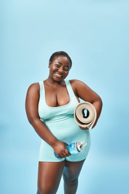 Neşeli ve iri yarı Afrikalı Amerikalı kadın spor paspası ve mavide su şişesiyle duruyor.