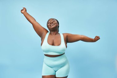 Mutlu artı boyut Afrikalı Amerikalı aktif kadın mavi arka planda neşeyle esniyor.