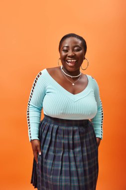 Heyecanlı ve Afrikalı Amerikan mankeni ekose etek ve nane rengi kazak giymiş turuncu üzerine poz veriyor.