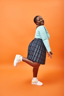 Ekose etek ve nane rengi kazakla turuncu üzerine poz veren Afro-Amerikan modeli.
