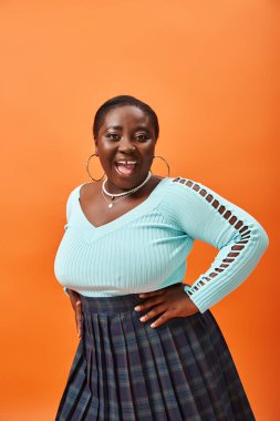 Ekoseli etek ve nane rengi uzun kollu turuncu poz veren Afro-Amerikan modeli.