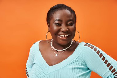 Neşeli, iri yarı, mavi kollu, turuncu arka planda poz veren Afro-Amerikan kadın.