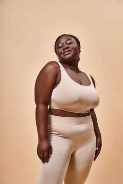 Işıl ışıl ve Afrikalı Amerikalı kadın bej rengi spor kıyafetleri içinde arka planda gülümsüyor.