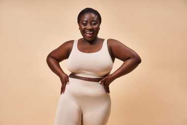 Radiant plus size african american woman in beige sportswear with joyful smile on matching backdrop clipart