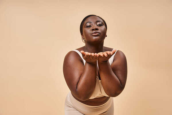 young plus size african american woman in beige underwear blowing air kiss against matching backdrop