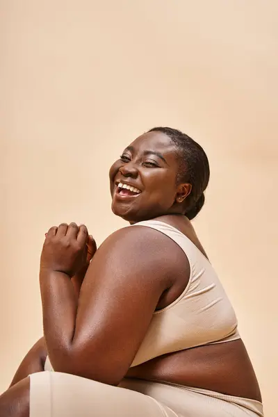 plus size african american model in beige lingerie laughing and reclining against matching backdrop