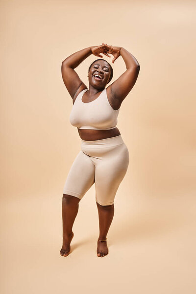 happy plus size woman in beige underwear posing with raised hands, body positive and self esteem