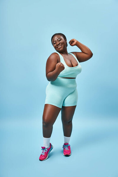 excited plus size woman in active wear flexing muscles and looking at camera on blue background