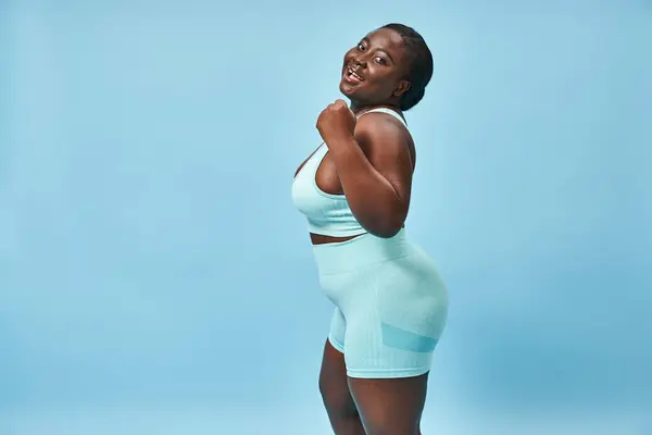 happy plus size woman in active wear flexing her muscles and looking at camera on blue background