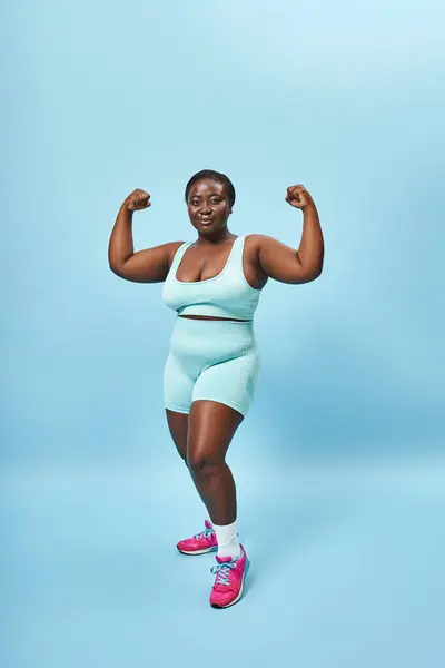 strong plus size woman in active wear flexing her muscles and looking at camera on blue background