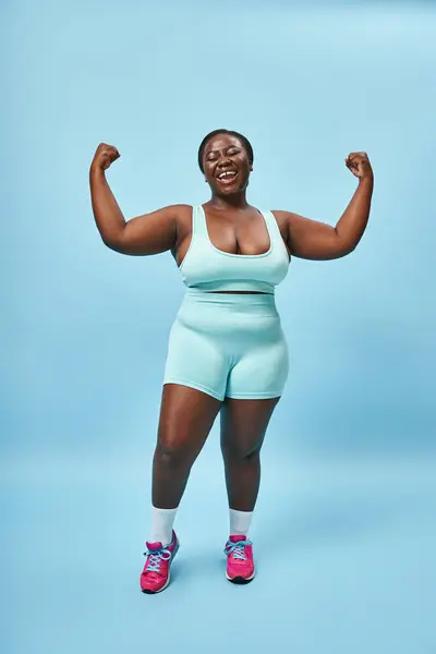excited plus size woman in active wear flexing her muscles and smiling on blue background