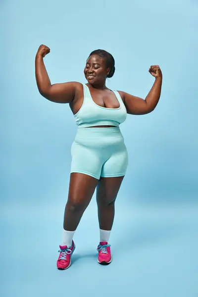 cheerful plus size woman in active wear flexing her muscles and smiling on blue background
