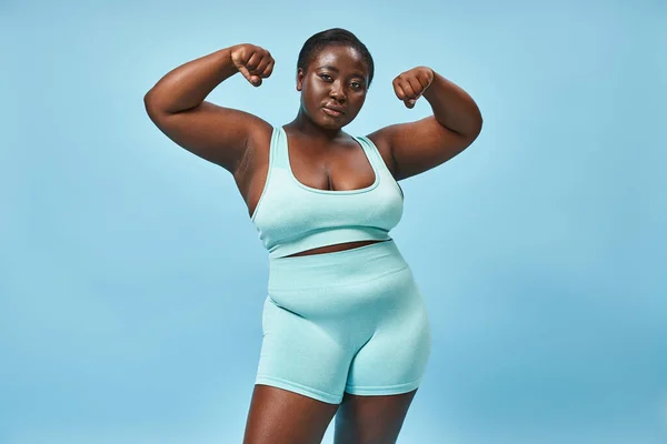 serious plus size woman in active wear flexing her muscles and looking at camera on blue background