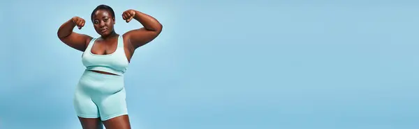 banner, plus size woman in active wear flexing her muscles and looking at camera on blue background