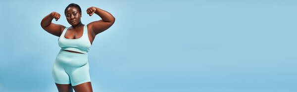 banner, plus size woman in active wear flexing her muscles and looking at camera on blue background