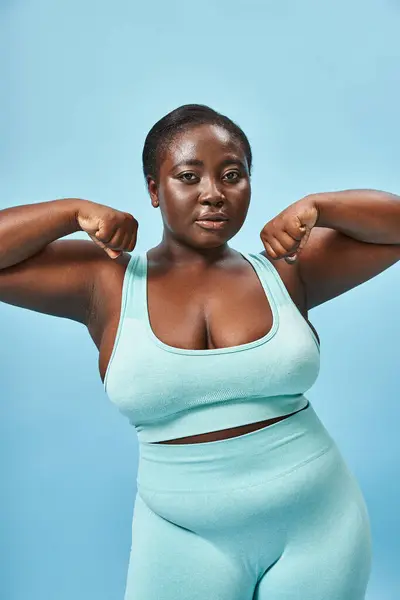 sport, plus size woman in active wear flexing her muscles and looking at camera on blue background
