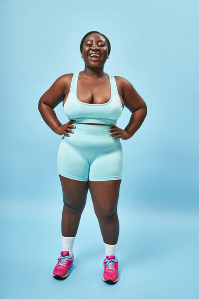 Весело плюс размер африканская американка в синей спортивной одежде с руками на бедрах на соответствующем фоне
